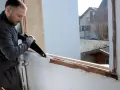 Un homme procède au démontage d'une fenêtre PVC avec une scie, révélant le cadre en bois sous-jacent.
