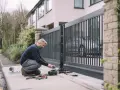 Un homme ajuste le moteur d'un portail automatique. Outils et pièces sont disposés sur le trottoir, prêts pour l'installation.