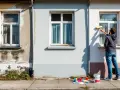 Un homme peint une fenêtre extérieure. Des pots de peinture et des nuanciers sont posés sur le trottoir, prêts pour la décoration de fenêtre extérieur.