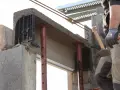 Un maçon prépare à installer une fenêtre dans un mur en béton, avec des étais soutenant un linteau fraîchement coulé.