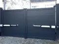 Portail gris à deux battants avec motorisation. Les bras de la motorisation portail sont fixés sur un mur perpendiculaire.