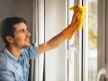 Un homme nettoie une fenêtre en aluminium avec un chiffon jaune. Le soleil brille à travers la vitre.