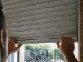 Un homme essaie de remonter son volet roulant qui ne fonctionne plus après coupure de courant. Il tient le bas du volet avec ses deux mains.