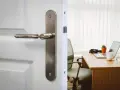 Poignée de porte en métal sur une porte blanche, révélant un bureau avec un ordinateur portable. La serrure BCC, un gage de sécurité.