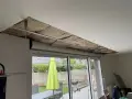 Travaux d'isolation phonique fenêtre PVC coffre : le plafond est ouvert, révélant l'isolant. Dehors, un jardin avec parasol vert.