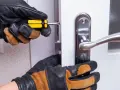 Mains gantées ajustent un loquet serrure bloqué sur une porte blanche avec un tournevis.