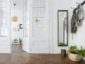 Un couloir lumineux avec une porte vitrée blanche ouverte, un miroir, des manteaux sur un porte-manteau et des plantes. Idéal pour un projet de relooker porte.