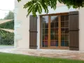 Fenêtre en bois avec volets bruns, matériaux menuiserie de qualité. Le jardin verdoyant et la façade en pierre complètent le tableau.