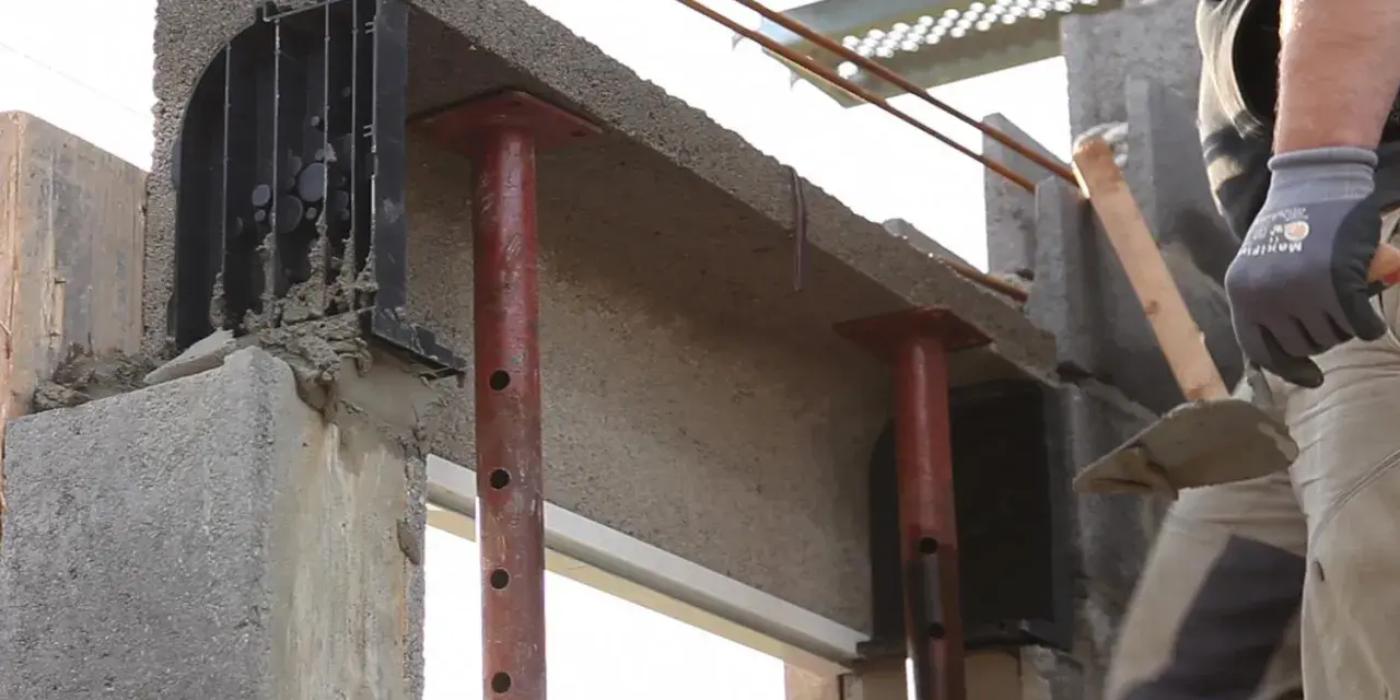 Un maçon prépare à installer une fenêtre dans un mur en béton, avec des étais soutenant un linteau fraîchement coulé.