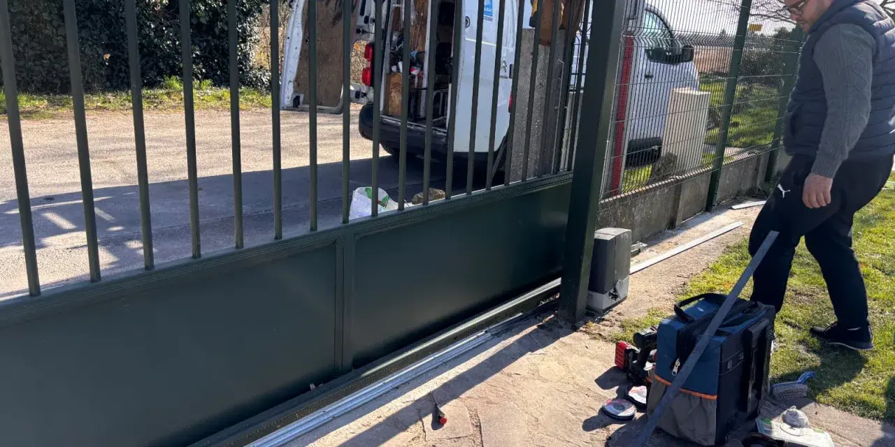 Un homme installe le moteur d'un portail coulissant. Des outils sont posés au sol près de la cellule.