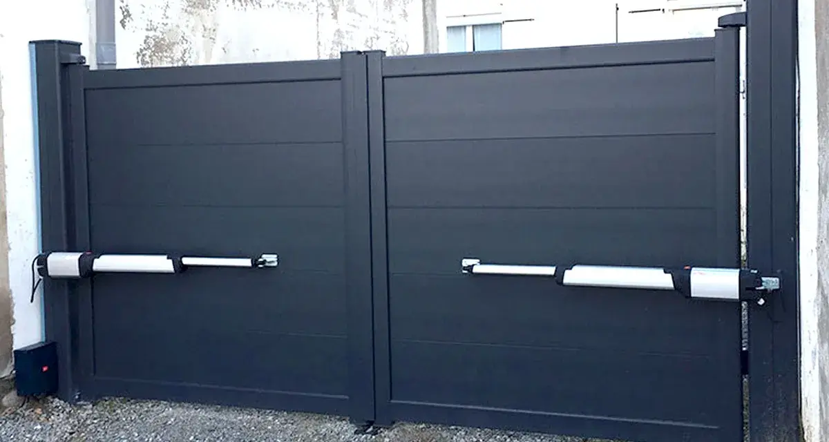 Portail gris à deux battants avec motorisation. Les bras de la motorisation portail sont fixés sur un mur perpendiculaire.
