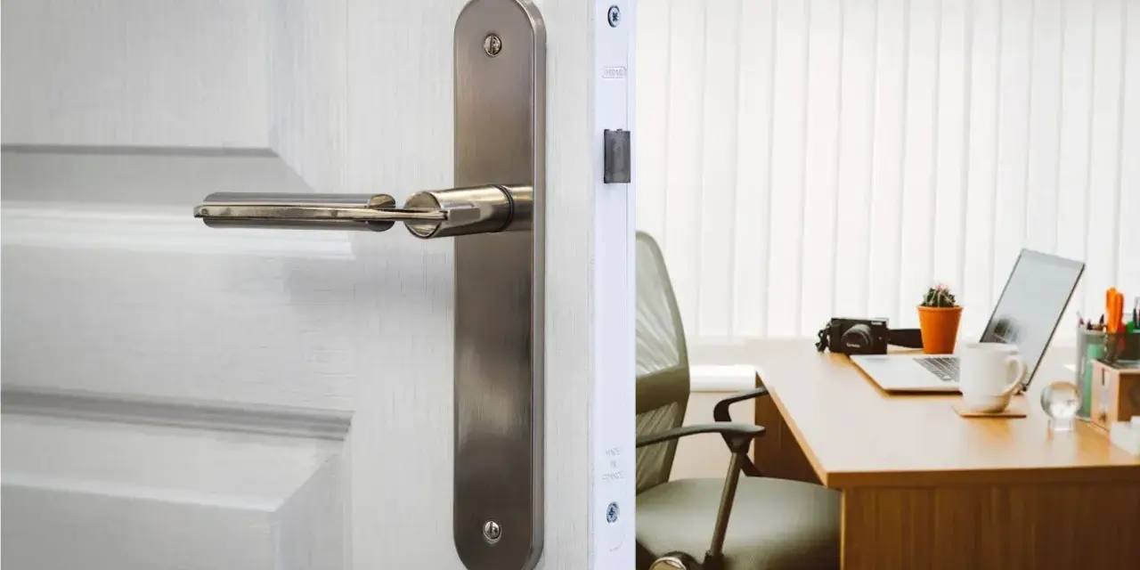 Poignée de porte en métal sur une porte blanche, révélant un bureau avec un ordinateur portable. La serrure BCC, un gage de sécurité.