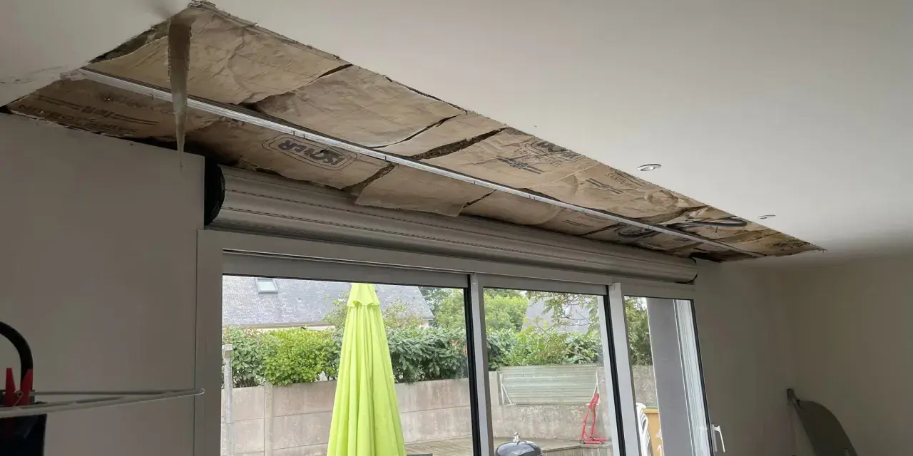 Travaux d'isolation phonique fenêtre PVC coffre : le plafond est ouvert, révélant l'isolant. Dehors, un jardin avec parasol vert.