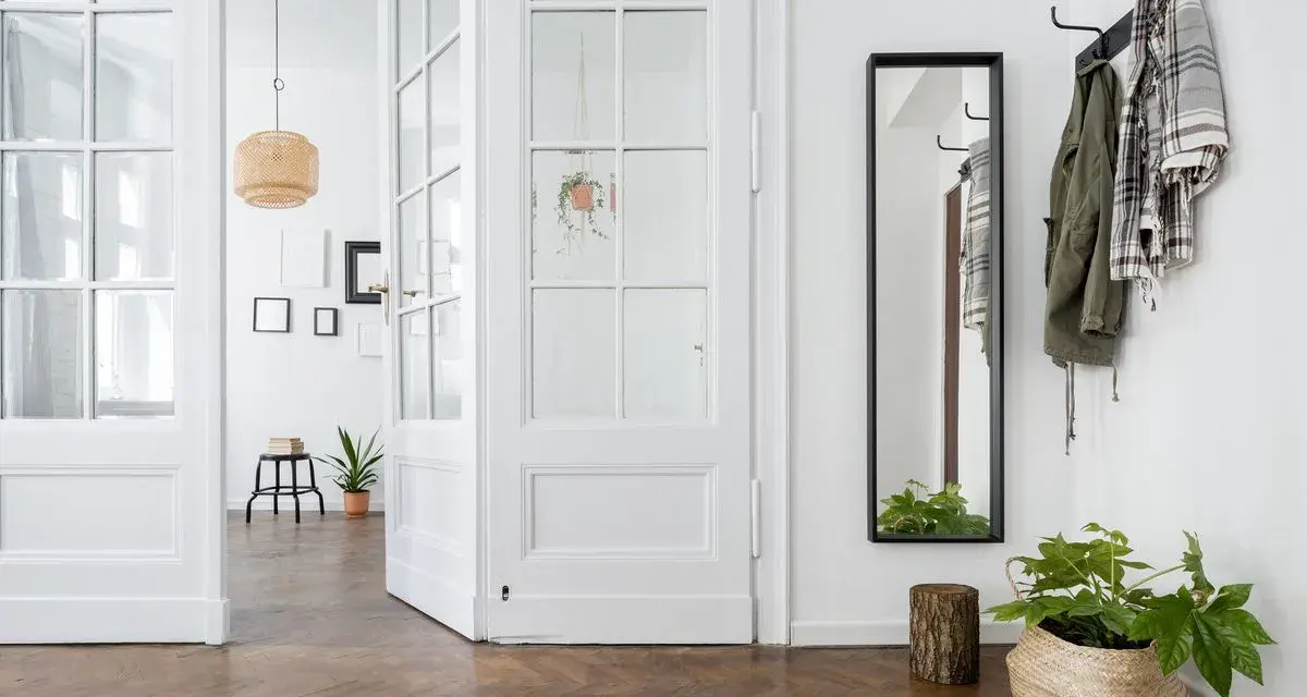 Un couloir lumineux avec une porte vitrée blanche ouverte, un miroir, des manteaux sur un porte-manteau et des plantes. Idéal pour un projet de relooker porte.