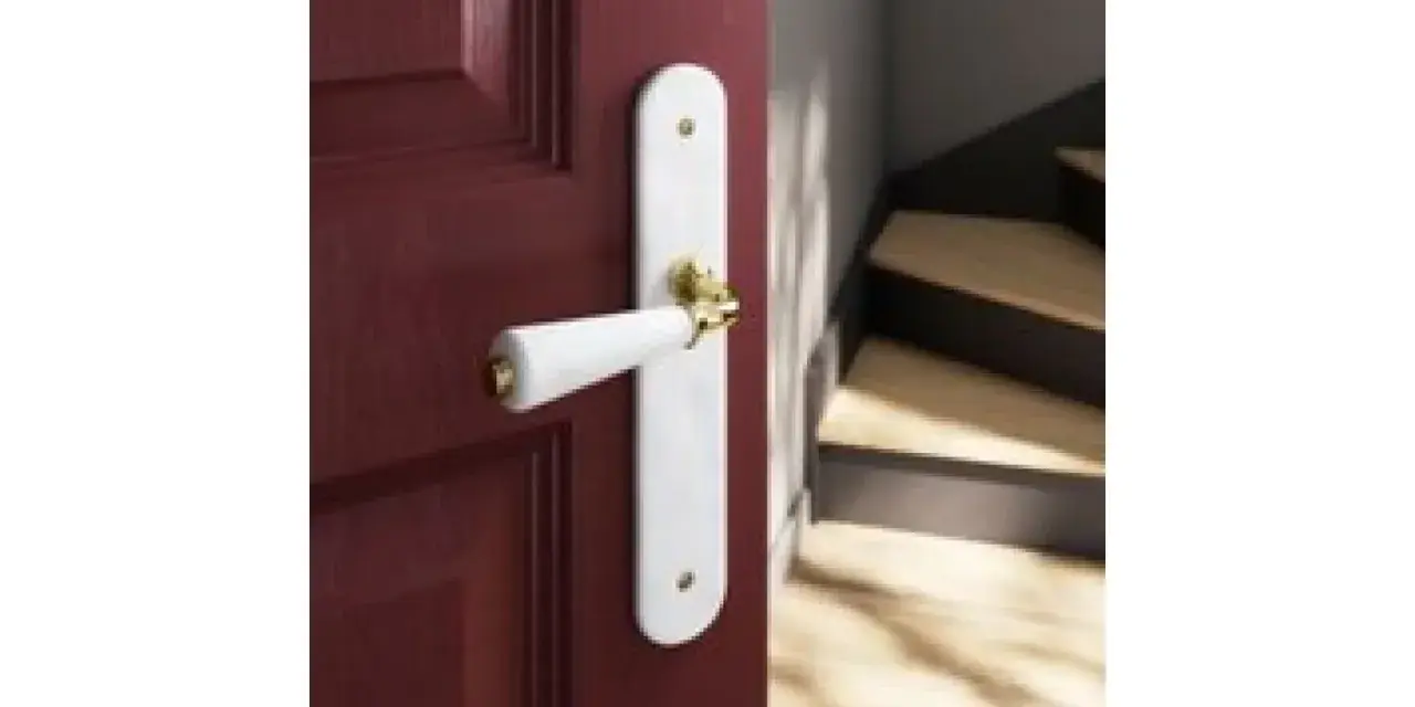 Poignée de porte blanche sur fond bordeaux, avec un escalier en bois visible. Quelle couleur de poignée pour porte blanche ? L'or brille.
