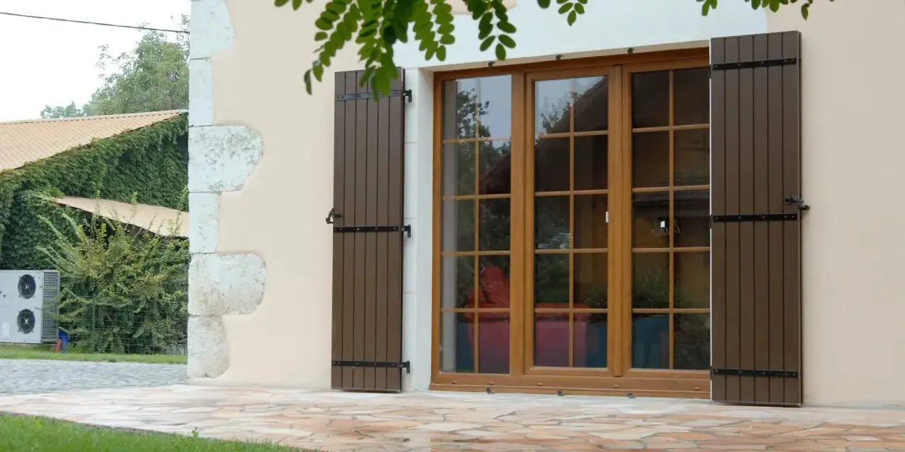 Fenêtre en bois avec volets bruns, matériaux menuiserie de qualité. Le jardin verdoyant et la façade en pierre complètent le tableau.