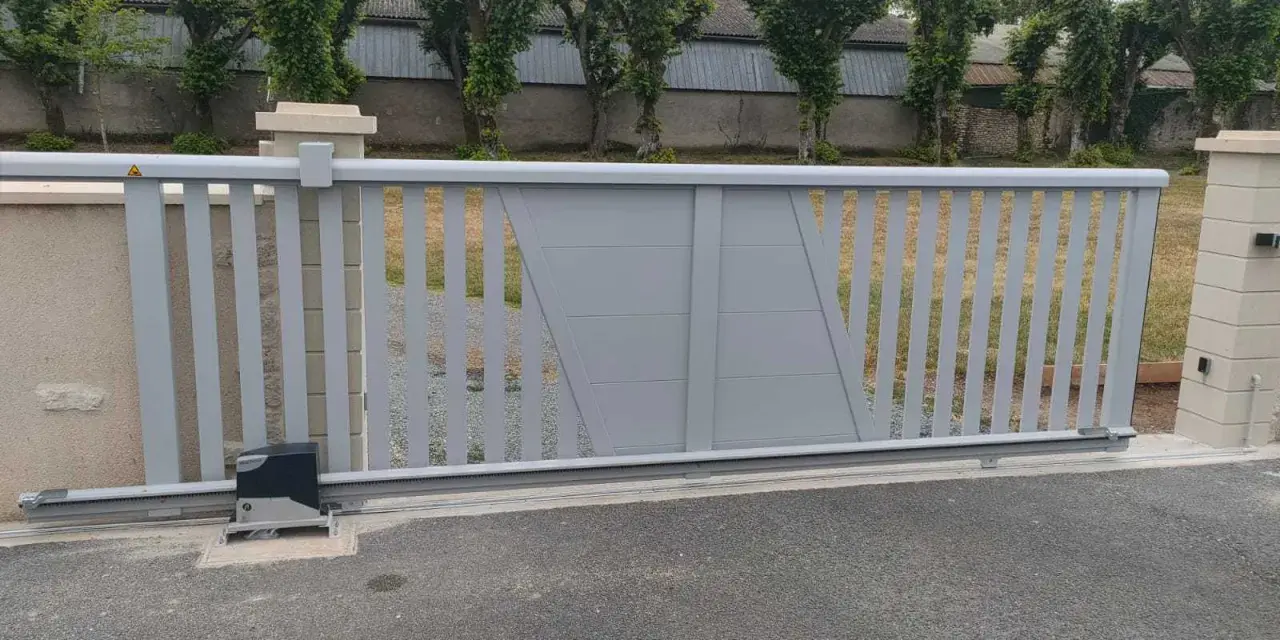 Portail électrique gris coulissant, avec son moteur discret, assure la protection du domicile. Des arbres taillés bordent la propriété.