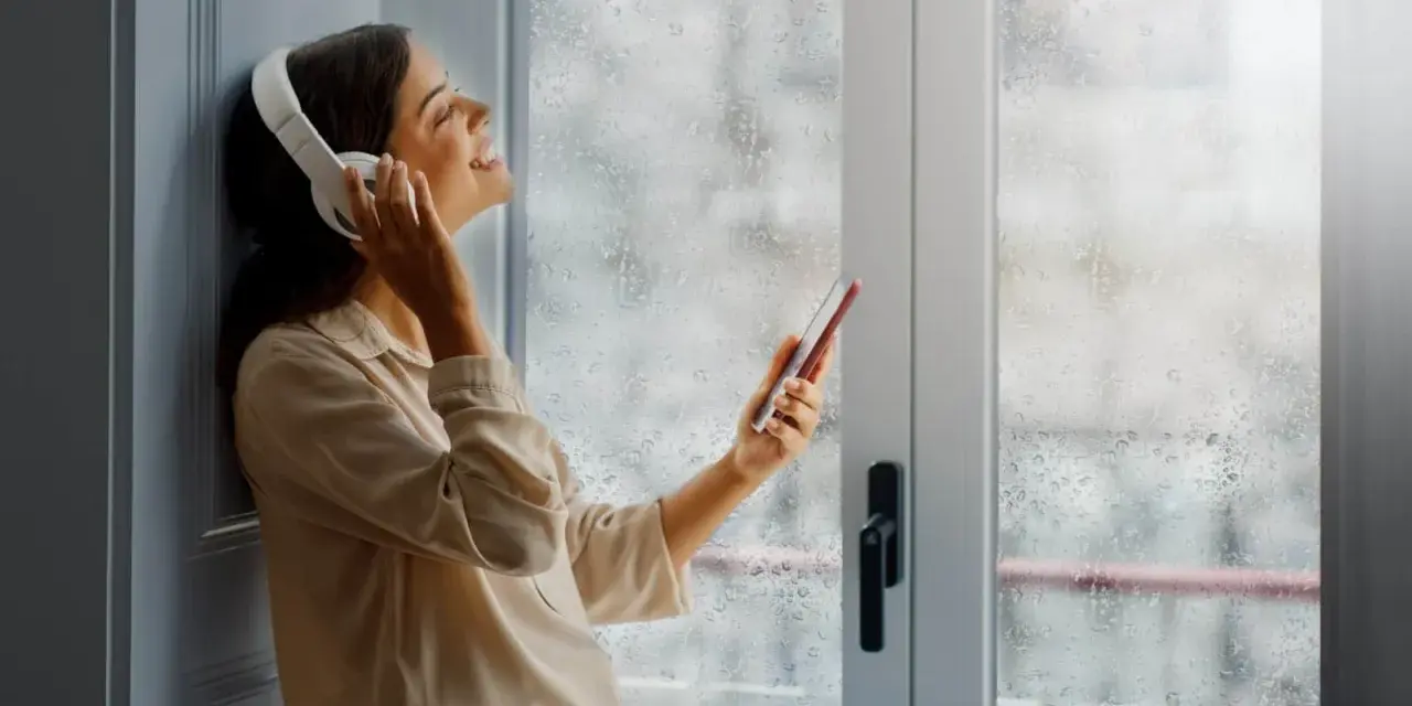 Une femme écoute de la musique avec des écouteurs, profitant du calme malgré la pluie. Un film isolant phonique pour fenêtre pourrait améliorer cette quiétude.