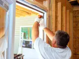 Un ouvrier installe une porte d'entrée, assurant une isolation parfaite. La structure en bois et les fils électriques sont visibles.