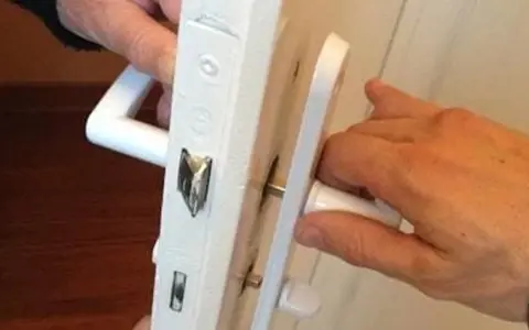 Deux mains actionnent une poignée de porte qui grince, révélant le mécanisme de serrure blanc.