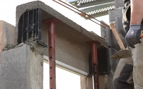 Un maçon prépare à installer une fenêtre dans un mur en béton, avec des étais soutenant un linteau fraîchement coulé.