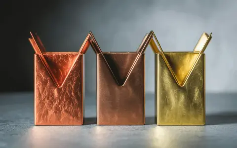 Trois porte-stylos aux formes géométriques, dont un en couleur laiton, sur un fond sombre.