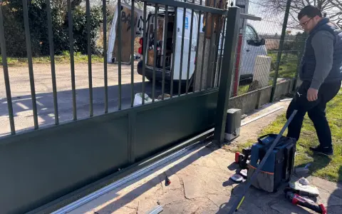 Un homme installe le moteur d'un portail coulissant. Des outils sont posés au sol près de la cellule.