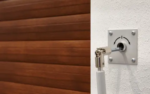 Mécanisme pour comment ouvrir une porte de garage électrique de l'extérieur. Un levier est inséré dans une serrure sur un mur blanc, à côté d'une porte en bois.