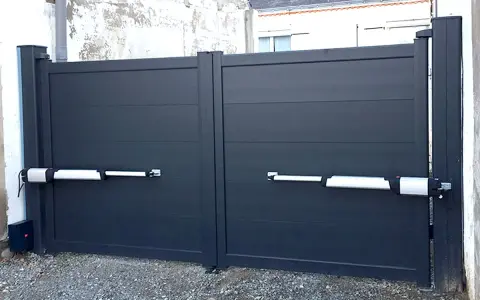 Portail gris à deux battants avec motorisation. Les bras de la motorisation portail sont fixés sur un mur perpendiculaire.