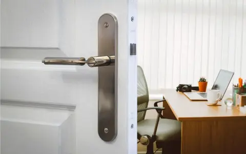 Poignée de porte en métal sur une porte blanche, révélant un bureau avec un ordinateur portable. La serrure BCC, un gage de sécurité.