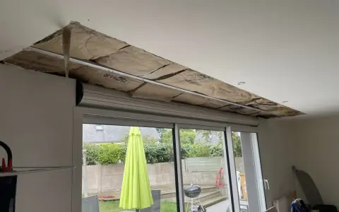 Travaux d'isolation phonique fenêtre PVC coffre : le plafond est ouvert, révélant l'isolant. Dehors, un jardin avec parasol vert.