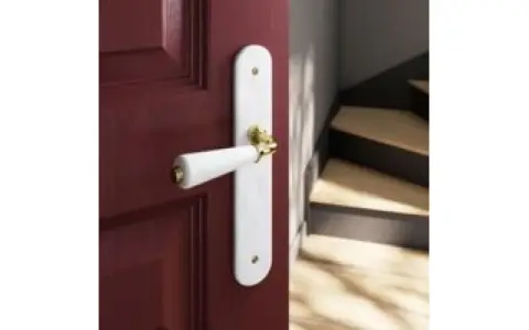Poignée de porte blanche sur fond bordeaux, avec un escalier en bois visible. Quelle couleur de poignée pour porte blanche ? L'or brille.