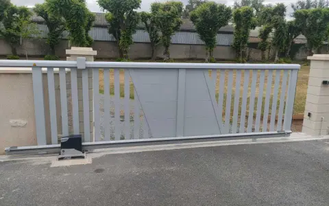 Portail électrique gris coulissant, avec son moteur discret, assure la protection du domicile. Des arbres taillés bordent la propriété.