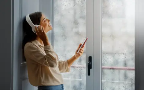 Une femme écoute de la musique avec des écouteurs, profitant du calme malgré la pluie. Un film isolant phonique pour fenêtre pourrait améliorer cette quiétude.