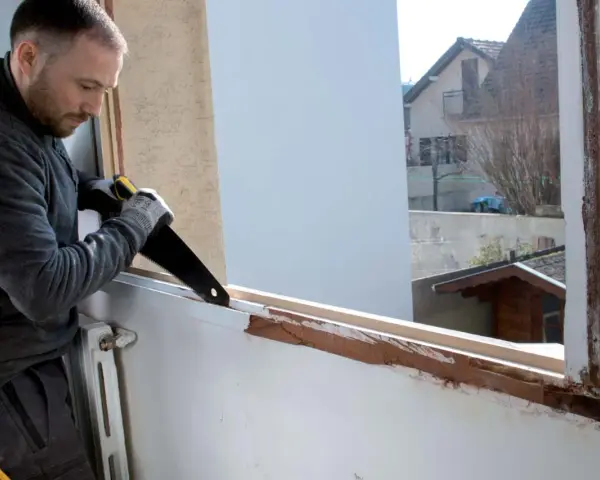 Un homme procède au démontage d'une fenêtre PVC avec une scie, révélant le cadre en bois sous-jacent.
