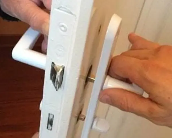 Deux mains actionnent une poignée de porte qui grince, révélant le mécanisme de serrure blanc.