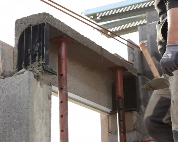 Un maçon prépare à installer une fenêtre dans un mur en béton, avec des étais soutenant un linteau fraîchement coulé.