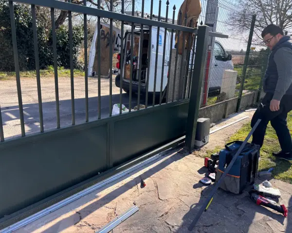 Un homme installe le moteur d'un portail coulissant. Des outils sont posés au sol près de la cellule.