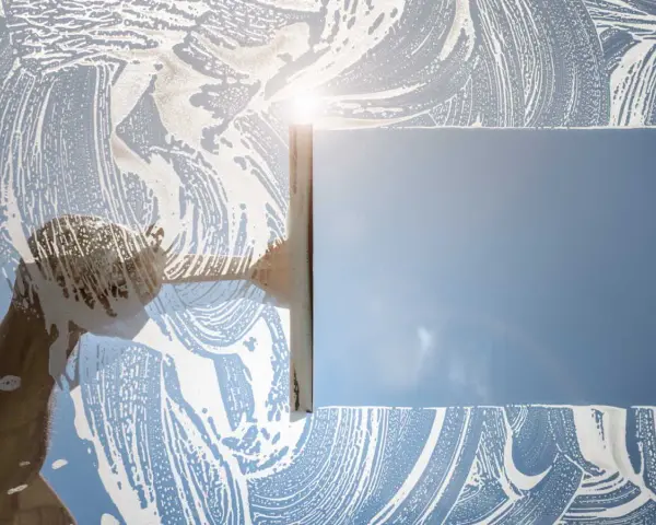 Nettoyage d'une vitre avec une raclette et de la mousse. Le soleil brille à travers la surface savonneuse, créant des reflets. Idéal pour enlever la colle sur vitre.