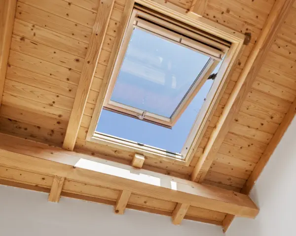 Un *uw velux* ouvert sur un ciel bleu éclatant, encadré par des poutres et un plafond en bois chaleureux.
