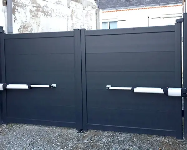 Portail gris à deux battants avec motorisation. Les bras de la motorisation portail sont fixés sur un mur perpendiculaire.
