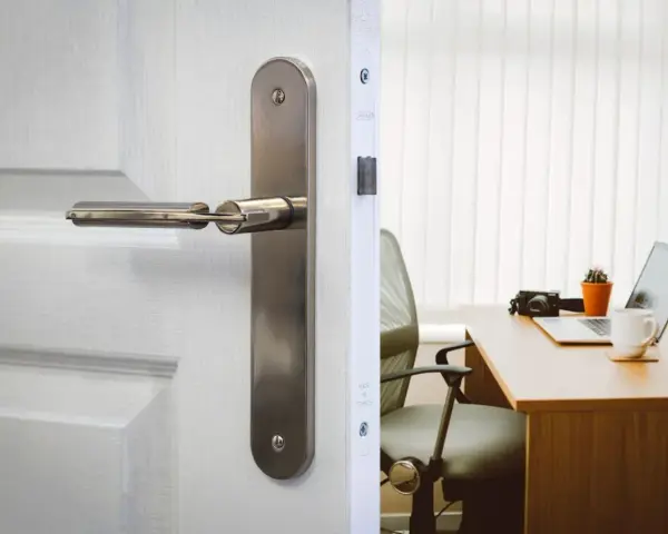 Poignée de porte en métal sur une porte blanche, révélant un bureau avec un ordinateur portable. La serrure BCC, un gage de sécurité.