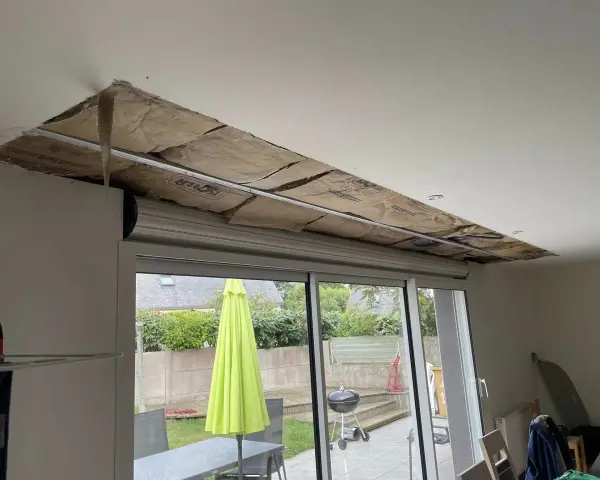 Travaux d'isolation phonique fenêtre PVC coffre : le plafond est ouvert, révélant l'isolant. Dehors, un jardin avec parasol vert.