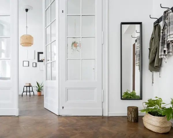 Un couloir lumineux avec une porte vitrée blanche ouverte, un miroir, des manteaux sur un porte-manteau et des plantes. Idéal pour un projet de relooker porte.