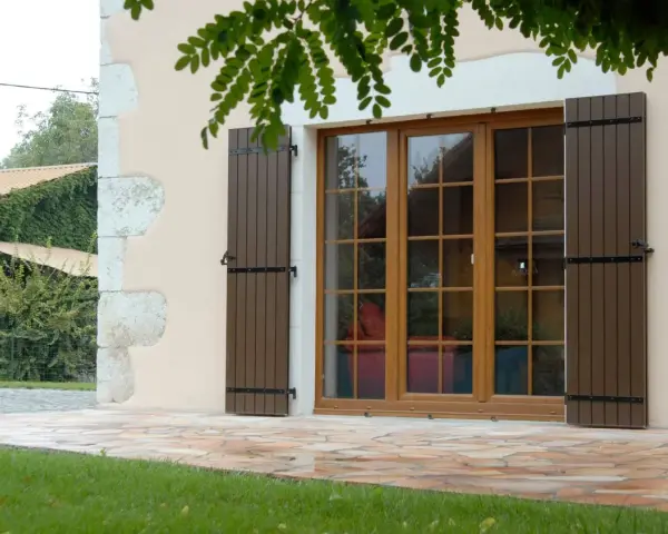 Fenêtre en bois avec volets bruns, matériaux menuiserie de qualité. Le jardin verdoyant et la façade en pierre complètent le tableau.