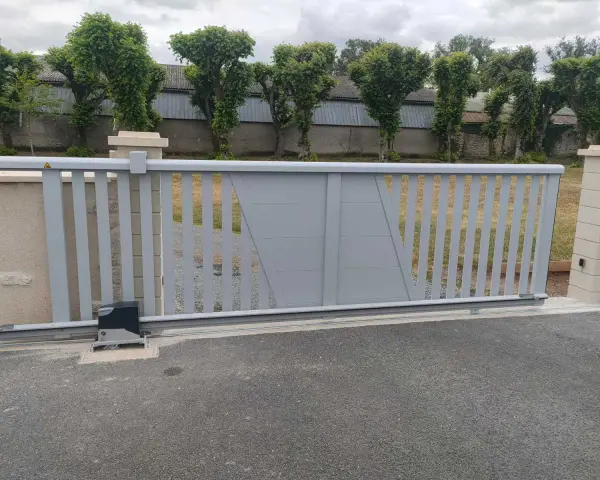 Portail électrique gris coulissant, avec son moteur discret, assure la protection du domicile. Des arbres taillés bordent la propriété.