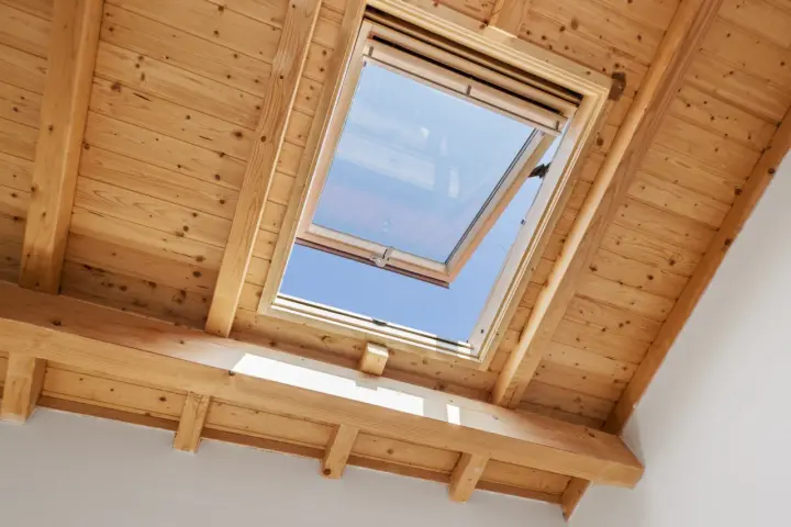 Un *uw velux* ouvert sur un ciel bleu éclatant, encadré par des poutres et un plafond en bois chaleureux.