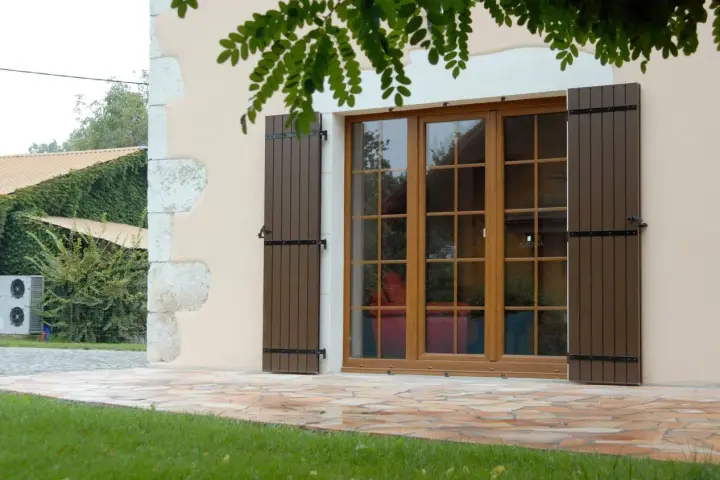 Fenêtre en bois avec volets bruns, matériaux menuiserie de qualité. Le jardin verdoyant et la façade en pierre complètent le tableau.