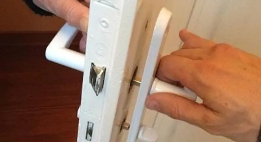 Deux mains actionnent une poignée de porte qui grince, révélant le mécanisme de serrure blanc.
