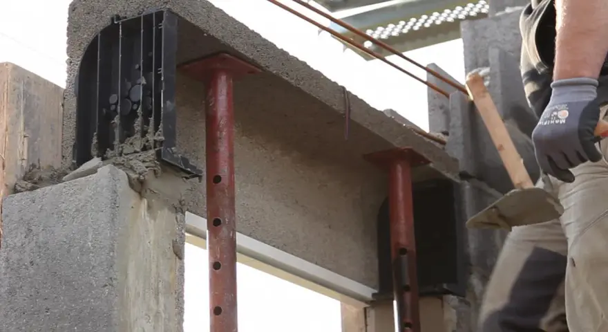 Un maçon prépare à installer une fenêtre dans un mur en béton, avec des étais soutenant un linteau fraîchement coulé.